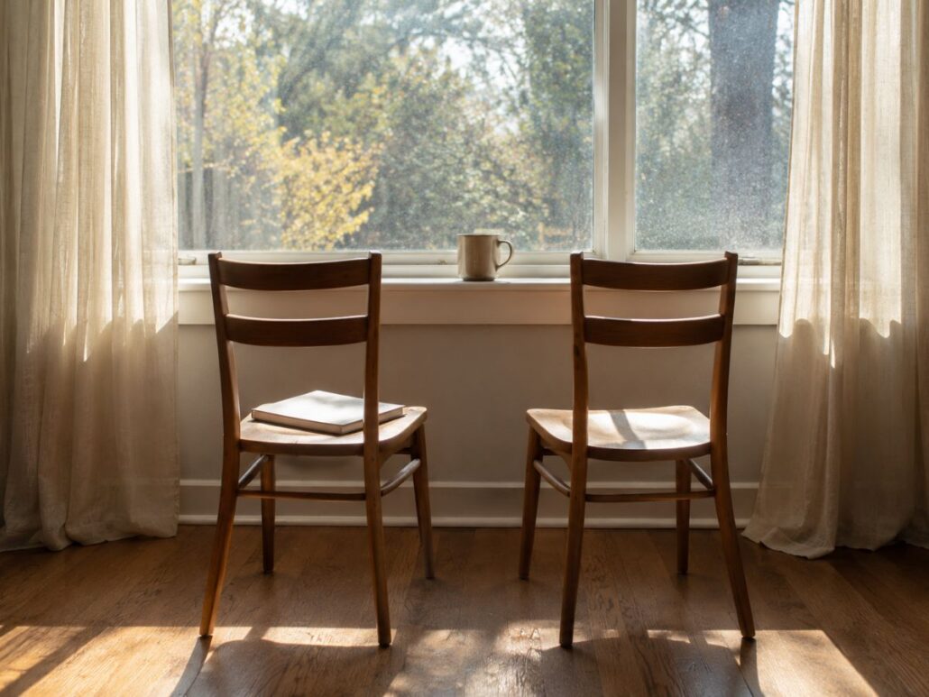 Two empty chairs side by side facing a bright window in a quiet room, with a mug on the sill.